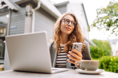 Genç bir kadın elinde telefon ve dizüstü bilgisayarla verandadaki bir masada oturuyor. Bir iş kadını kahve içer ve dışarıda dizüstü bilgisayarla çalışır. Serbest ve kahve molası kavramı.