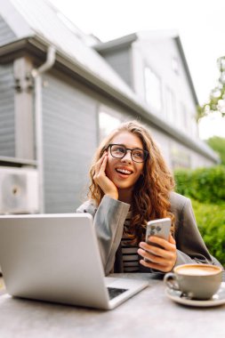 Genç bir kadın elinde telefon ve dizüstü bilgisayarla verandadaki bir masada oturuyor. Bir iş kadını kahve içer ve dışarıda dizüstü bilgisayarla çalışır. Serbest ve kahve molası kavramı.