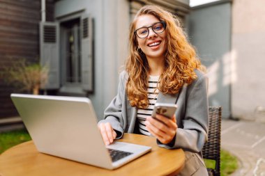 Genç bir kadın elinde telefon ve dizüstü bilgisayarla verandadaki bir masada oturuyor. Bir iş kadını kahve içer ve dışarıda dizüstü bilgisayarla çalışır. Serbest ve kahve molası kavramı.