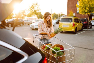 Taze ürünlerle dolu çantalarla gülümseyen kadın gün batımında alışverişini arabasının bagajına koyuyor. Güneşli bir otoparkta, arabasının yanında market arabasıyla güzel bir kadın. Tüketim kavramı.