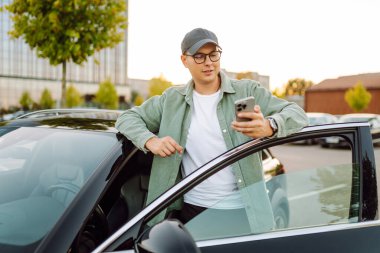 Gözlüklü ve şapkalı genç bir adam güneşli bir otoparkta arabanın yanında duruyor. Şık bir araba sürücüsü otoparkta durmuş sohbet ediyor. Teknoloji ve ulaşım kavramı.