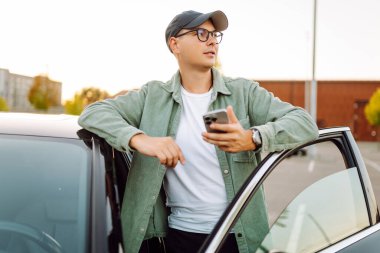 Gözlüklü ve şapkalı genç bir adam güneşli bir otoparkta arabanın yanında duruyor. Şık bir araba sürücüsü otoparkta durmuş sohbet ediyor. Teknoloji ve ulaşım kavramı.