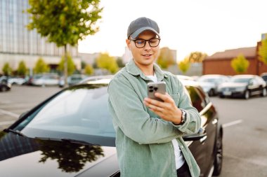 Gözlüklü ve şapkalı genç bir adam güneşli bir otoparkta arabanın yanında duruyor. Şık bir araba sürücüsü otoparkta durmuş sohbet ediyor. Teknoloji ve ulaşım kavramı.
