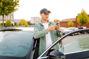 Gözlüklü ve şapkalı genç bir adam güneşli bir otoparkta arabanın yanında duruyor. Şık bir araba sürücüsü otoparkta durmuş sohbet ediyor. Teknoloji ve ulaşım kavramı.