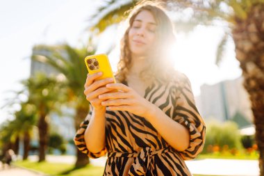 Güneşli bir caddede duran genç bir kadının elindeki parlak bir çantadaki telefonun yakın çekimi. Genç turist dışarıda telefonuyla mesajlaşıyor ve blog yazıyor. Turizm ve eğlence konsepti.