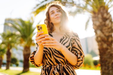 Güneşli bir caddede duran genç bir kadının elindeki parlak bir çantadaki telefonun yakın çekimi. Genç turist dışarıda telefonuyla mesajlaşıyor ve blog yazıyor. Turizm ve eğlence konsepti.