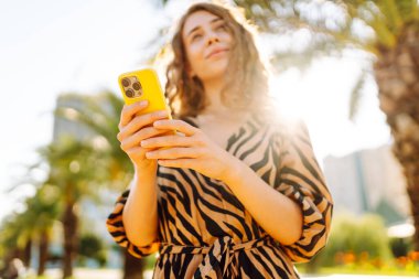 Güneşli bir caddede duran genç bir kadının elindeki parlak bir çantadaki telefonun yakın çekimi. Genç turist dışarıda telefonuyla mesajlaşıyor ve blog yazıyor. Turizm ve eğlence konsepti.