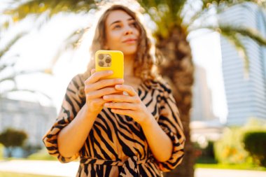 Güneşli bir caddede duran genç bir kadının elindeki parlak bir çantadaki telefonun yakın çekimi. Genç turist dışarıda telefonuyla mesajlaşıyor ve blog yazıyor. Turizm ve eğlence konsepti.