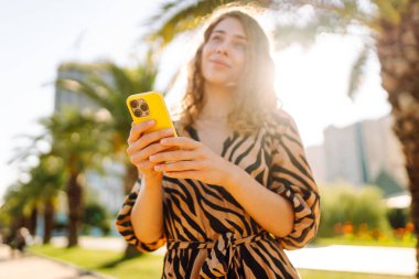 Güneşli bir caddede duran genç bir kadının elindeki parlak bir çantadaki telefonun yakın çekimi. Genç turist dışarıda telefonuyla mesajlaşıyor ve blog yazıyor. Turizm ve eğlence konsepti.