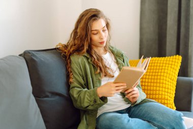 Gözlüklü genç bir kadın rahat bir odada kanepede kitap okumaktan zevk alıyor. Gözlüklü hoş bir kadın kitap okuyor, evde çalışıyor ve dinleniyor. Eğitim, rahatlama ve rahatlama konsepti.
