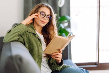 Gözlüklü genç bir kadın rahat bir odada kanepede kitap okumaktan zevk alıyor. Gözlüklü hoş bir kadın kitap okuyor, evde çalışıyor ve dinleniyor. Eğitim, rahatlama ve rahatlama konsepti.