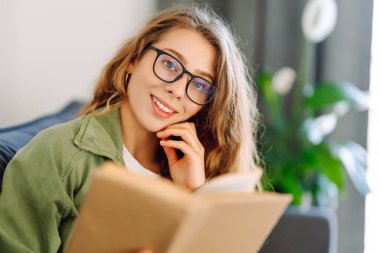 Gözlüklü genç bir kadın rahat bir odada kanepede kitap okumaktan zevk alıyor. Gözlüklü hoş bir kadın kitap okuyor, evde çalışıyor ve dinleniyor. Eğitim, rahatlama ve rahatlama konsepti.