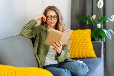 Gözlüklü genç bir kadın rahat bir odada kanepede kitap okumaktan zevk alıyor. Gözlüklü hoş bir kadın kitap okuyor, evde çalışıyor ve dinleniyor. Eğitim, rahatlama ve rahatlama konsepti.