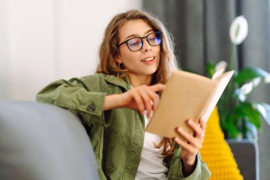 Gözlüklü genç bir kadın rahat bir odada kanepede kitap okumaktan zevk alıyor. Gözlüklü hoş bir kadın kitap okuyor, evde çalışıyor ve dinleniyor. Eğitim, rahatlama ve rahatlama konsepti.