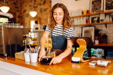 Barın arkasında elinde bir fincan sıcak ve aromatik kahve tutan mutlu bayan barista. Kahve dükkanının genç çalışanı bir müşteriye kahve verir. İçki kavramı, küçük işletmeler..