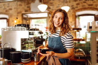 Barın arkasında elinde bir fincan sıcak ve aromatik kahve tutan mutlu bayan barista. Kahve dükkanının genç çalışanı bir müşteriye kahve verir. İçki kavramı, küçük işletmeler..