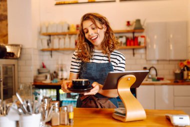 Barın arkasında elinde bir fincan sıcak ve aromatik kahve tutan mutlu bayan barista. Kahve dükkanının genç çalışanı bir müşteriye kahve verir. İçki kavramı, küçük işletmeler..