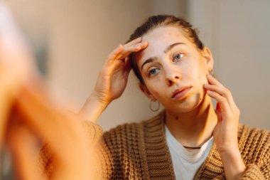 Aynaya bakarken cildine döküntülerle dokunan genç bir kadının portresi. Rahat bir odada sorunlu tenli güzel bir kadın. Kozmetik kavramı, doğal güzellik.