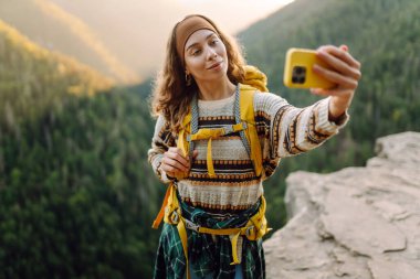 Parlak sırt çantalı mutlu bir kadının portresi kayalık bir çıkıntıda selfie çeker. Genç gezgin blogları ya da modern bir telefonla fotoğraf çekiyor ve dağ manzarasının tadını çıkarıyor. Doğa konsepti.