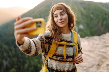Parlak sırt çantalı mutlu bir kadının portresi kayalık bir çıkıntıda selfie çeker. Genç gezgin blogları ya da modern bir telefonla fotoğraf çekiyor ve dağ manzarasının tadını çıkarıyor. Doğa konsepti.