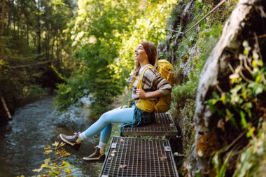 Sırt çantalı gülümseyen bir kadın sık güneşli ormanda kayalık yolda yürüyor. Genç bayan yürüyüşçü dağlık alanda yürüyüş yapıyor. Özgürlük, macera ve doğa kavramı.
