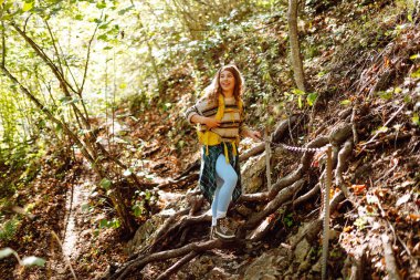Sırt çantalı gülümseyen bir kadın sık güneşli ormanda kayalık yolda yürüyor. Genç bayan yürüyüşçü dağlık alanda yürüyüş yapıyor. Özgürlük, macera ve doğa kavramı.