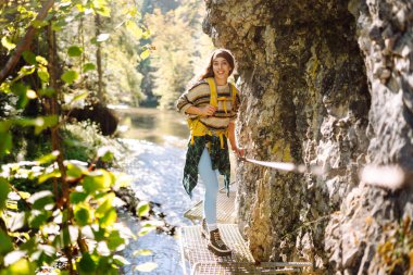 Sırt çantalı gülümseyen bir kadın sık güneşli ormanda kayalık yolda yürüyor. Genç bayan yürüyüşçü dağlık alanda yürüyüş yapıyor. Özgürlük, macera ve doğa kavramı.