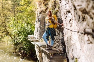 Sırt çantalı gülümseyen bir kadın sık güneşli ormanda kayalık yolda yürüyor. Genç bayan yürüyüşçü dağlık alanda yürüyüş yapıyor. Özgürlük, macera ve doğa kavramı.