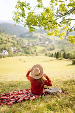 Yeşil bir tepenin üstündeki battaniyenin üzerinde oturan şapkalı genç bir kadının arka görüntüsü. Güzel bayan gezgin tepe manzarasının tadını çıkarıyor ve doğada özgürlüğü hissediyor. Seyahat kavramı.