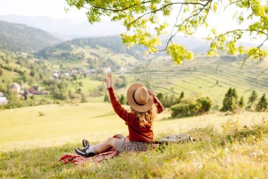 Yeşil bir tepenin üstündeki battaniyenin üzerinde oturan şapkalı genç bir kadının arka görüntüsü. Güzel bayan gezgin tepe manzarasının tadını çıkarıyor ve doğada özgürlüğü hissediyor. Seyahat kavramı.