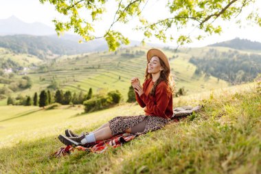 Yeşil bir tepenin üstündeki battaniyenin üzerinde oturan şapkalı genç bir kadının arka görüntüsü. Güzel bayan gezgin tepe manzarasının tadını çıkarıyor ve doğada özgürlüğü hissediyor. Seyahat kavramı.