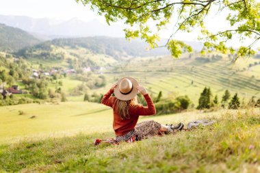 Yeşil bir tepenin üstündeki battaniyenin üzerinde oturan şapkalı genç bir kadının arka görüntüsü. Güzel bayan gezgin tepe manzarasının tadını çıkarıyor ve doğada özgürlüğü hissediyor. Seyahat kavramı.