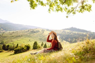 Yeşil bir tepenin üstündeki battaniyenin üzerinde oturan şapkalı genç bir kadının arka görüntüsü. Güzel bayan gezgin tepe manzarasının tadını çıkarıyor ve doğada özgürlüğü hissediyor. Seyahat kavramı.