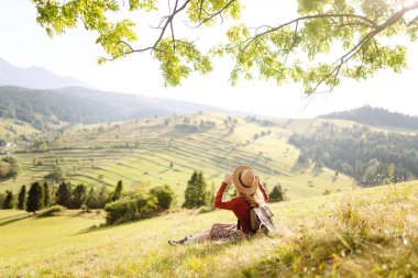 Yeşil bir tepenin üstündeki battaniyenin üzerinde oturan şapkalı genç bir kadının arka görüntüsü. Güzel bayan gezgin tepe manzarasının tadını çıkarıyor ve doğada özgürlüğü hissediyor. Seyahat kavramı.