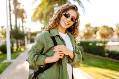 Kıvırcık saçlı ve güneş gözlüklü neşeli kadın palmiye ağaçlarıyla dolu bir sokakta güneşli bir günün tadını çıkarıyor. Sırt çantalı güzel bir turist dışarıda yürüyor. Boş zaman, hafta sonu konsepti..