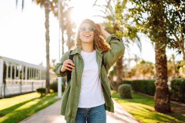 Kıvırcık saçlı ve güneş gözlüklü neşeli kadın palmiye ağaçlarıyla dolu bir sokakta güneşli bir günün tadını çıkarıyor. Sırt çantalı güzel bir turist dışarıda yürüyor. Boş zaman, hafta sonu konsepti..