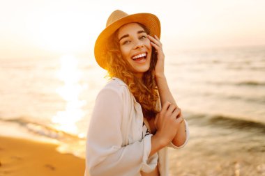 Şapkalı mutlu kadın gün batımında kumlu bir sahilde eğleniyor. Genç bayan deniz manzarasının tadını çıkarıyor ve dışarıda dinleniyor. Doğa, hafta sonu ve özgürlük kavramı.
