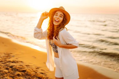 Şapkalı mutlu kadın gün batımında kumlu bir sahilde eğleniyor. Genç bayan deniz manzarasının tadını çıkarıyor ve dışarıda dinleniyor. Doğa, hafta sonu ve özgürlük kavramı.
