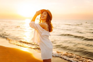 Şapkalı mutlu kadın gün batımında kumlu bir sahilde eğleniyor. Genç bayan deniz manzarasının tadını çıkarıyor ve dışarıda dinleniyor. Doğa, hafta sonu ve özgürlük kavramı.