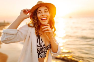 Şapkalı mutlu kadın gün batımında kumlu bir sahilde eğleniyor. Genç bayan deniz manzarasının tadını çıkarıyor ve dışarıda dinleniyor. Doğa, hafta sonu ve özgürlük kavramı.