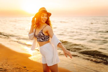 Şapkalı mutlu kadın gün batımında kumlu bir sahilde eğleniyor. Genç bayan deniz manzarasının tadını çıkarıyor ve dışarıda dinleniyor. Doğa, hafta sonu ve özgürlük kavramı.