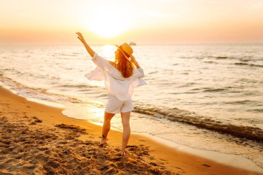 Şapkalı mutlu kadın gün batımında kumlu bir sahilde eğleniyor. Genç bayan deniz manzarasının tadını çıkarıyor ve dışarıda dinleniyor. Doğa, hafta sonu ve özgürlük kavramı.