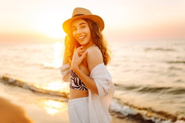 Şapkalı mutlu kadın gün batımında kumlu bir sahilde eğleniyor. Genç bayan deniz manzarasının tadını çıkarıyor ve dışarıda dinleniyor. Doğa, hafta sonu ve özgürlük kavramı.