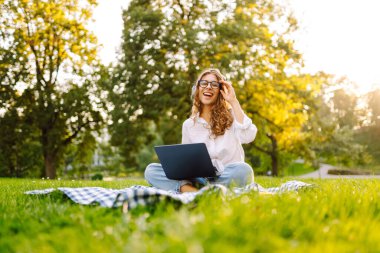 Neşeli kadın güneşli bir parkta dizüstü bilgisayarla battaniyeye oturur ve kablosuz kulaklıkla müzik dinler. Dışarıda telefonu ve dizüstü bilgisayarı olan güzel bir kadın çalışıyor. Serbest konsept, teknoloji.