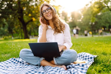 Neşeli kadın güneşli bir parkta dizüstü bilgisayarla battaniyeye oturur ve kablosuz kulaklıkla müzik dinler. Dışarıda telefonu ve dizüstü bilgisayarı olan güzel bir kadın çalışıyor. Serbest konsept, teknoloji.