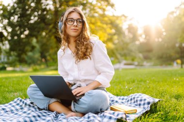 Neşeli kadın güneşli bir parkta dizüstü bilgisayarla battaniyeye oturur ve kablosuz kulaklıkla müzik dinler. Dışarıda telefonu ve dizüstü bilgisayarı olan güzel bir kadın çalışıyor. Serbest konsept, teknoloji.