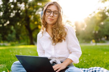 Neşeli kadın güneşli bir parkta dizüstü bilgisayarla battaniyeye oturur ve kablosuz kulaklıkla müzik dinler. Dışarıda telefonu ve dizüstü bilgisayarı olan güzel bir kadın çalışıyor. Serbest konsept, teknoloji.