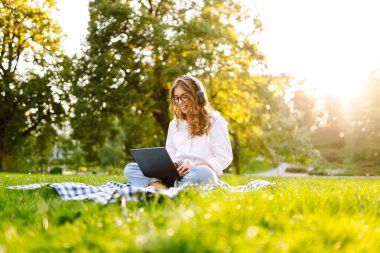Neşeli kadın güneşli bir parkta dizüstü bilgisayarla battaniyeye oturur ve kablosuz kulaklıkla müzik dinler. Dışarıda telefonu ve dizüstü bilgisayarı olan güzel bir kadın çalışıyor. Serbest konsept, teknoloji.