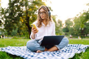 Neşeli kadın güneşli bir parkta dizüstü bilgisayarla battaniyeye oturur ve kablosuz kulaklıkla müzik dinler. Dışarıda telefonu ve dizüstü bilgisayarı olan güzel bir kadın çalışıyor. Serbest konsept, teknoloji.