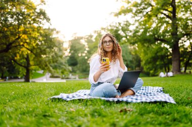 Neşeli kadın güneşli bir parkta dizüstü bilgisayarla battaniyeye oturur ve kablosuz kulaklıkla müzik dinler. Dışarıda telefonu ve dizüstü bilgisayarı olan güzel bir kadın çalışıyor. Serbest konsept, teknoloji.